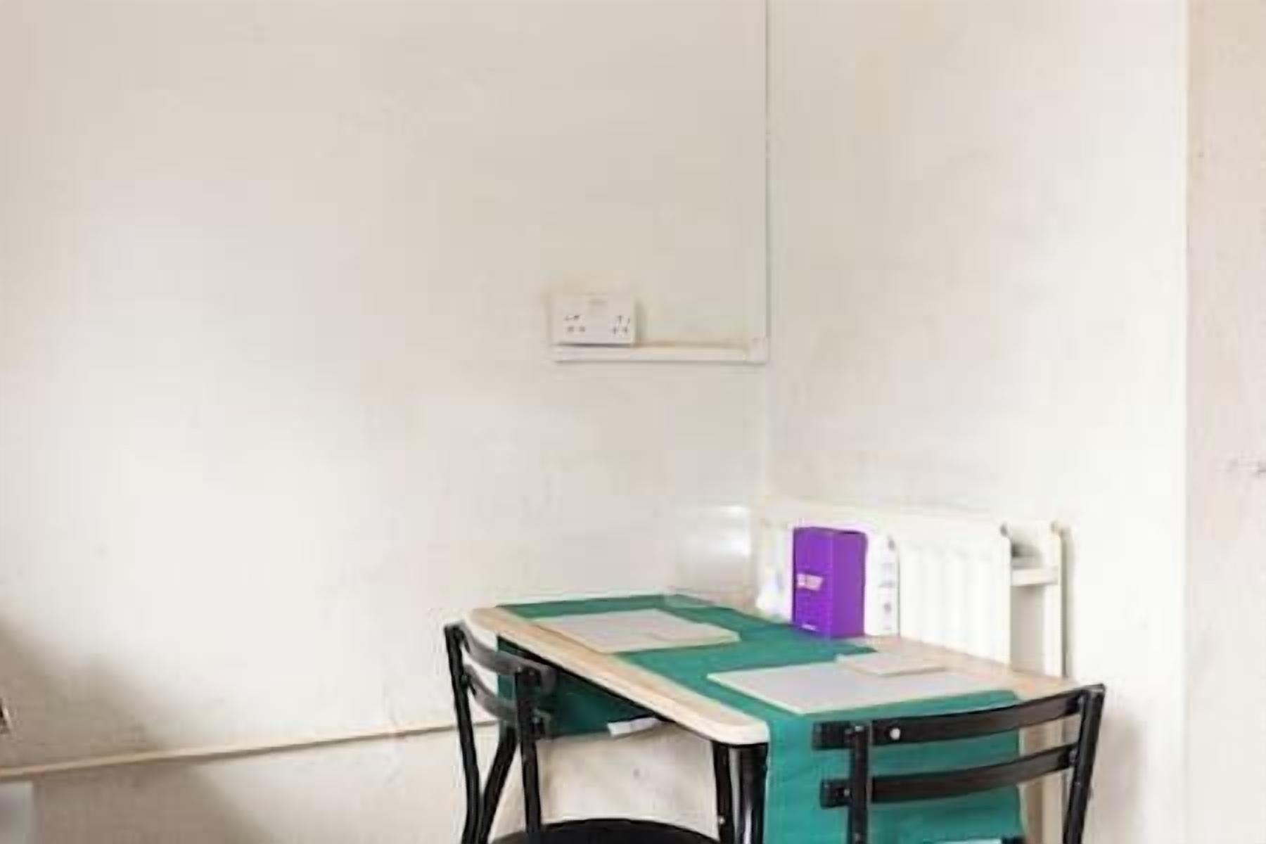 Una habitación pequeña y sencilla, con una mesa, una silla, un radiador blanco y una caja morada en la pared.