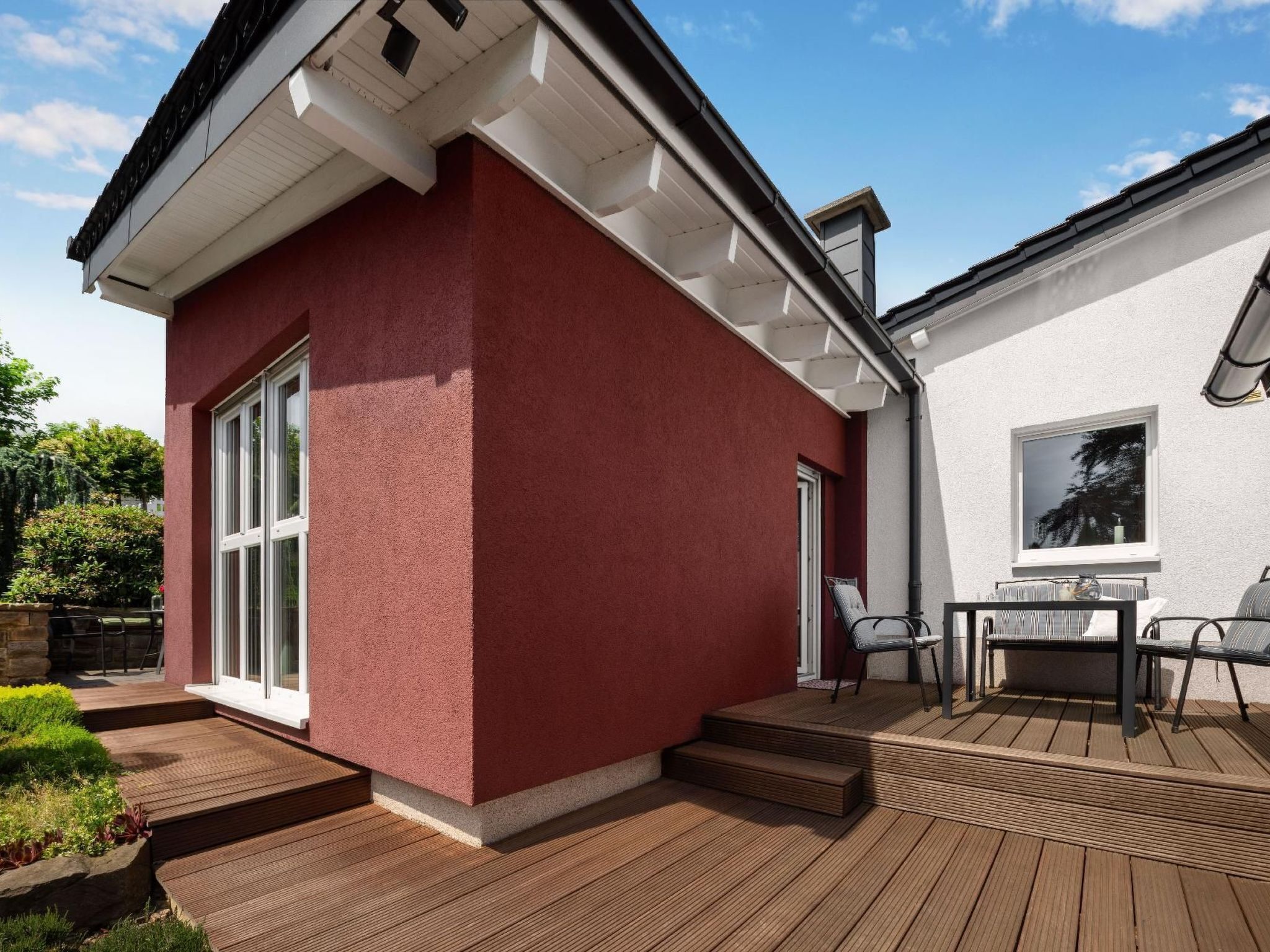 A modern house with a red exterior, large windows, and a deck with outdoor furniture.