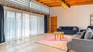Een ruime woonkamer met een houten plafond, een grote schuifpui, een blauwe bank en een grijze fauteuil.