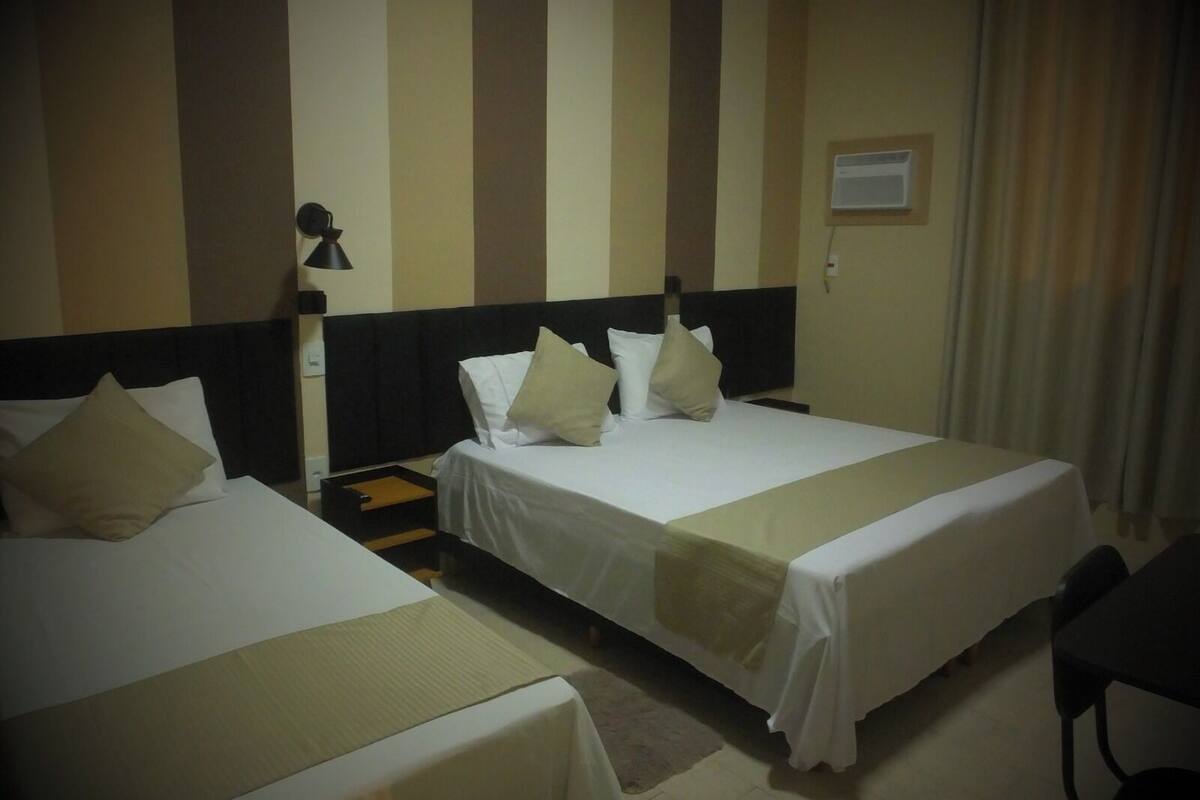 A hotel room with two beds, a nightstand, a lamp, and a window with curtains.