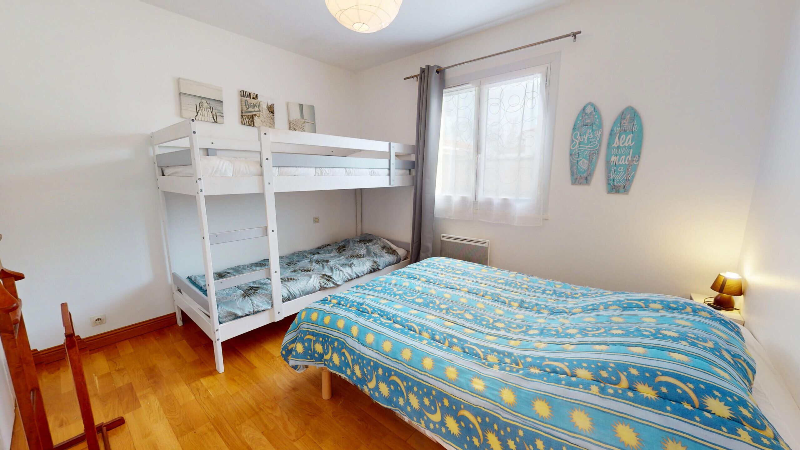 Une chambre avec un lit superposé, un lit simple avec une couvre-lit à motifs, une fenêtre avec des rideaux et deux planches de surf accrochées au mur.