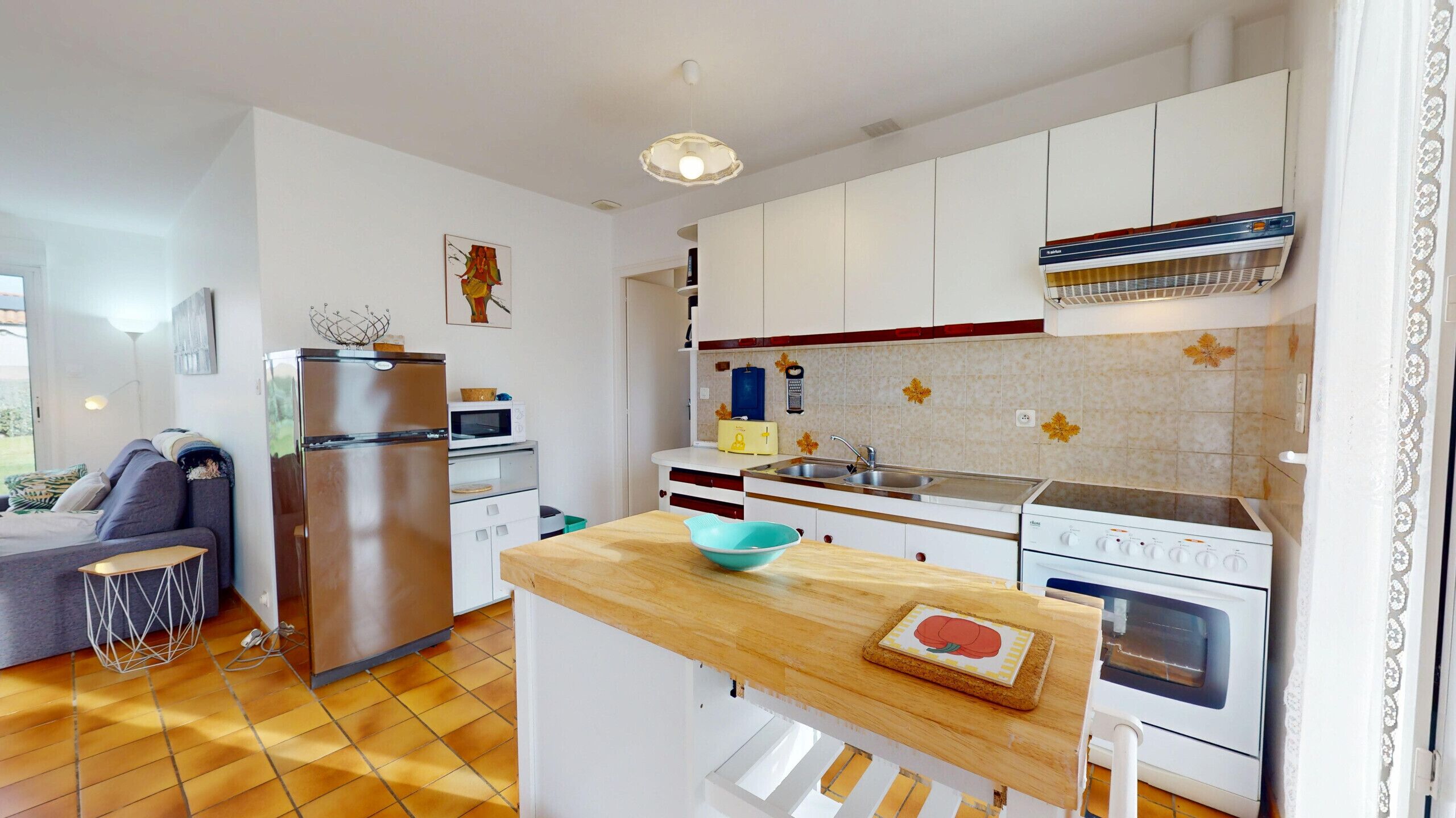 Une cuisine compacte avec des meubles blancs, un réfrigérateur en acier inoxydable, un four à micro-ondes et une cuisinière. On y trouve un plan de travail en bois, une planche à découper et un saladier.