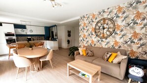 Ein modernes Wohnzimmer mit einem runden Esstisch, einer Couch mit Kissen, einem Couchtisch und einer Wand mit tropischem Blattmuster.