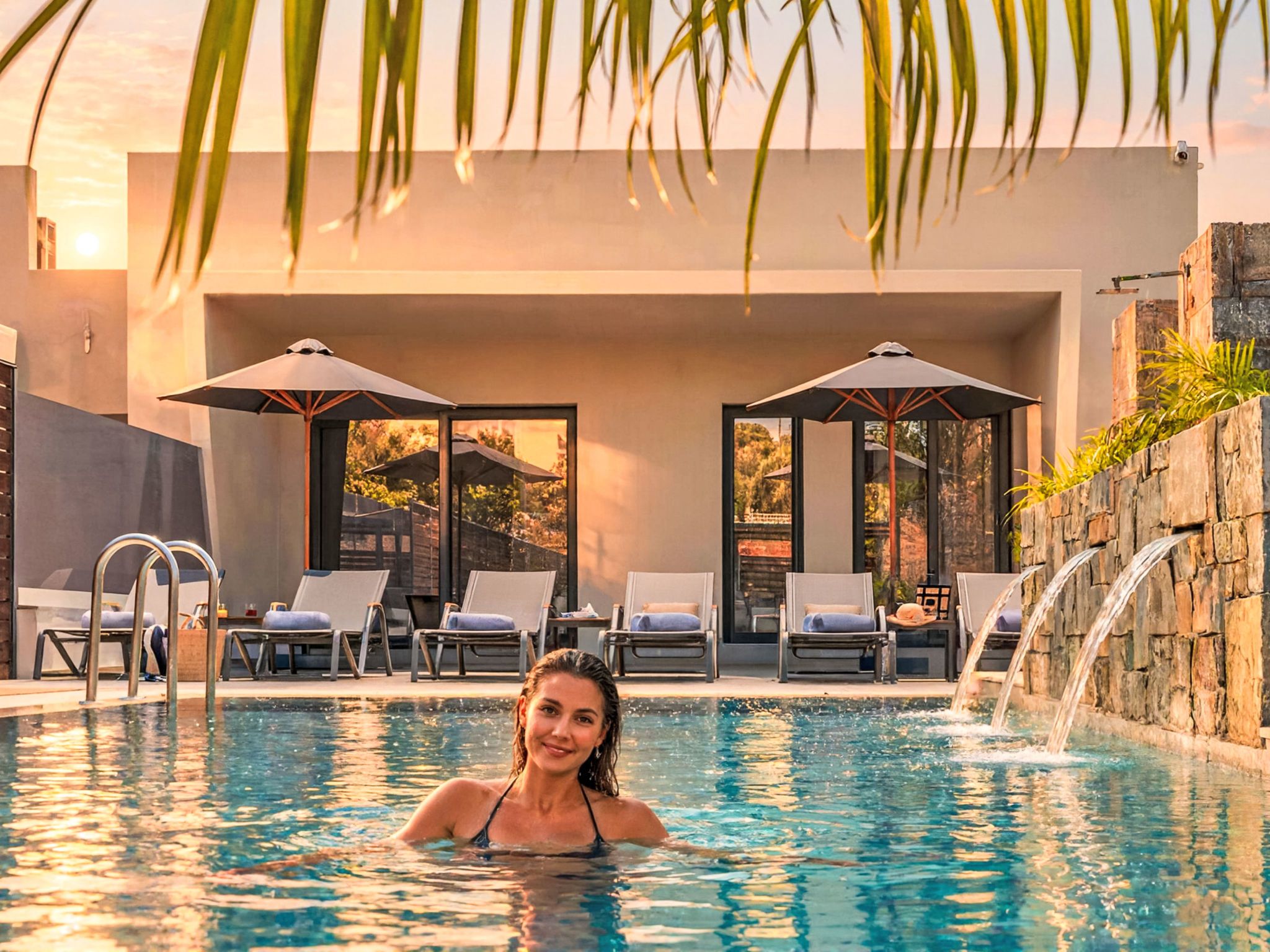 A swimming pool with lounge chairs and umbrellas, surrounded by stone walls and a view of a building.
