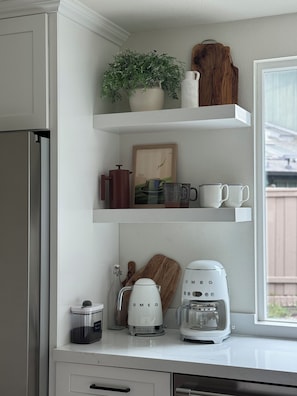 Una cocina con armarios blancos, cafetera, licuadora, hervidor y una planta en maceta sobre la encimera.