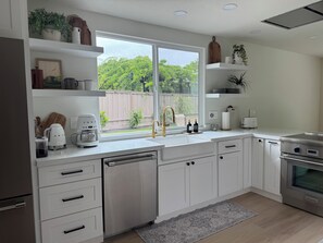 Una cocina moderna con armarios blancos, estufa de acero inoxidable, lavavajillas y ventana con vistas al verdor.