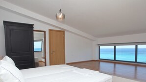 Une chambre avec un grand lit, une armoire, un miroir et une vue sur la mer grâce à de grandes fenêtres.