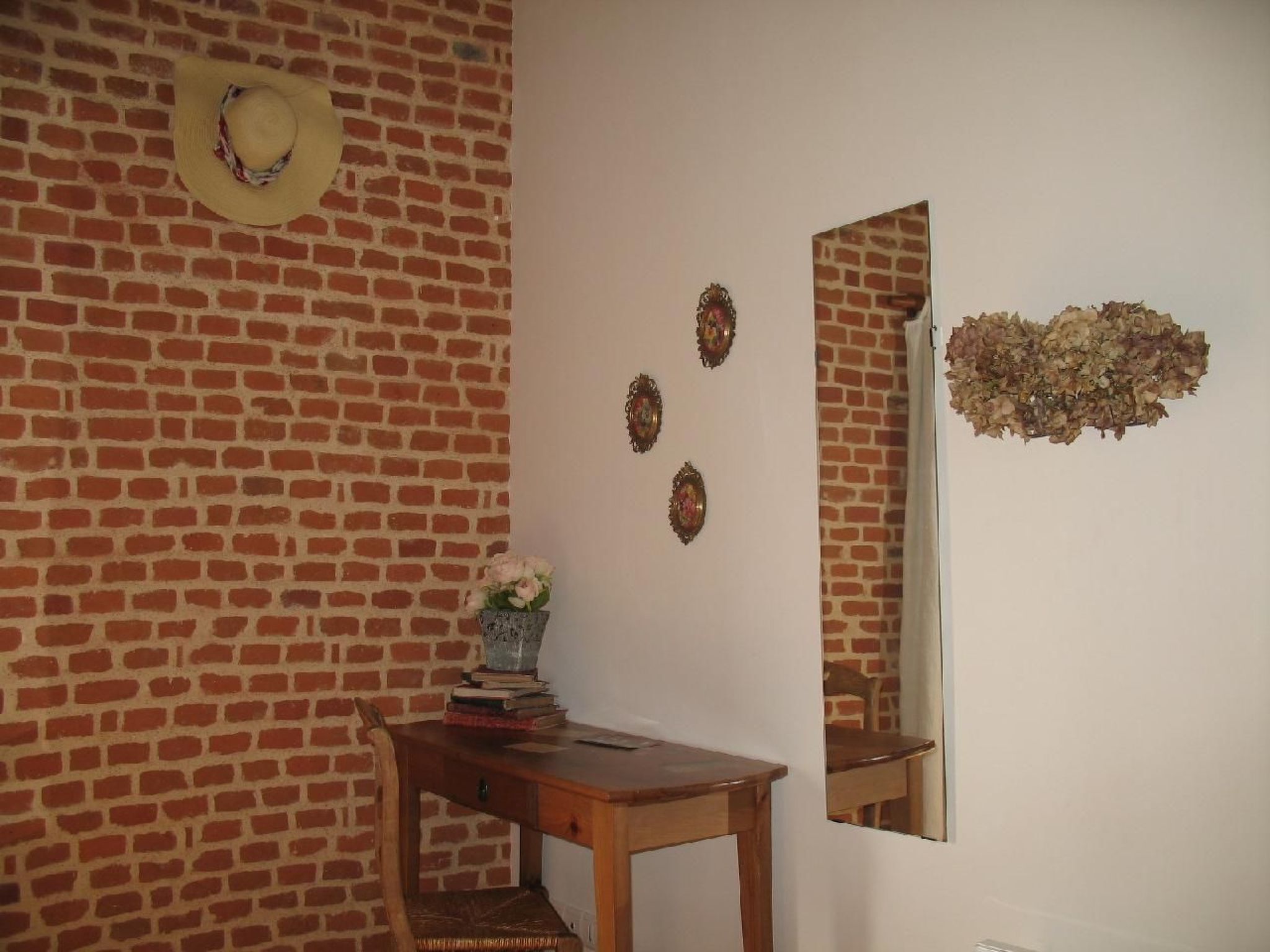 A room with a brick wall, a wooden desk with a vase of flowers, and a mirror reflecting the wall.