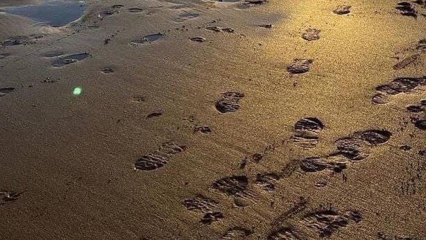Ein Strand mit Fußspuren und einem Leuchtturm in der Ferne.