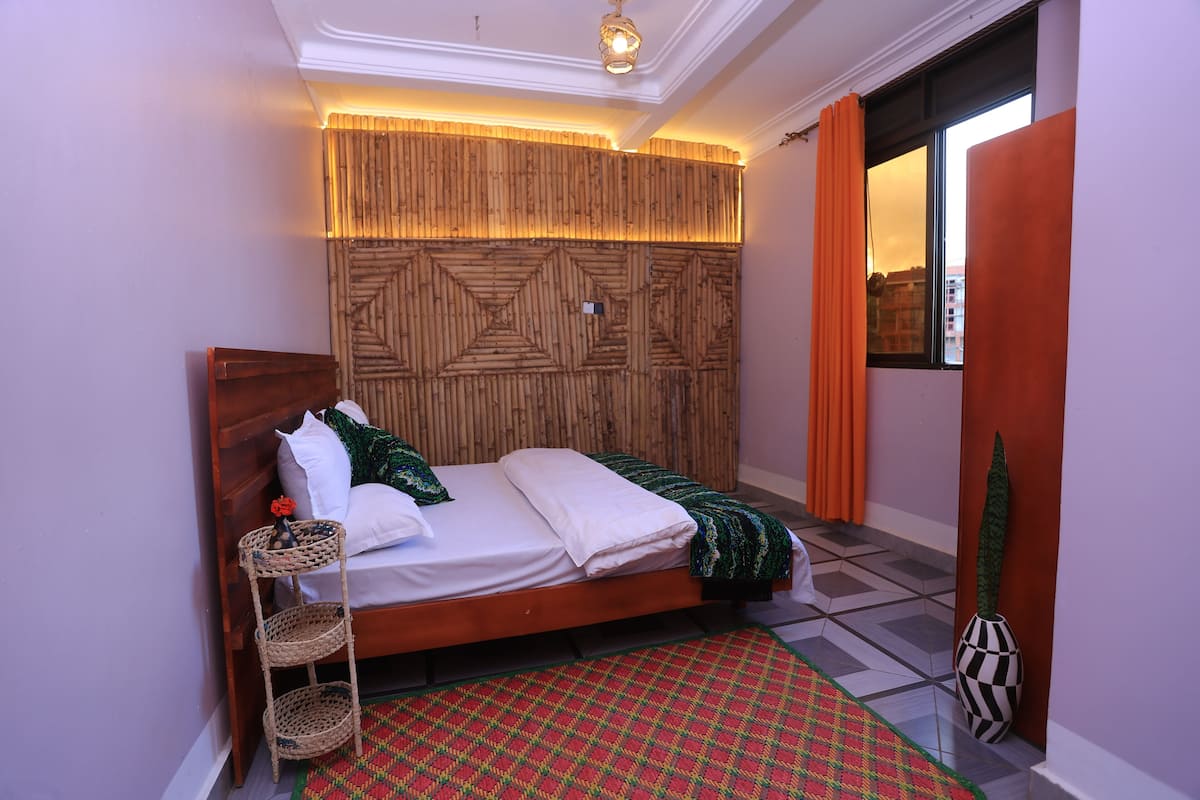 Un dormitorio con cama, cabecero fijado a la pared, mesita de noche, lámpara de pie y ventana con cortinas.