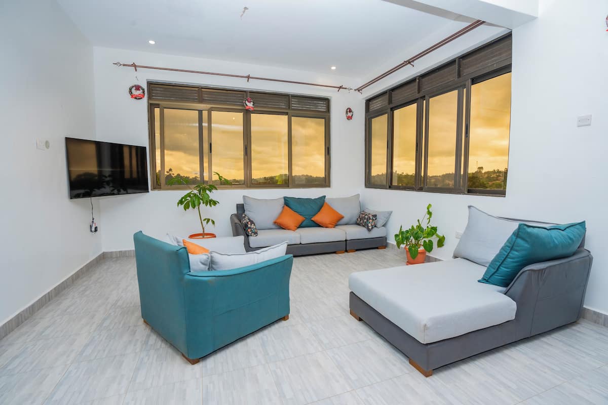Una sala de estar moderna con un sofá, un sillón y una mesa de centro. Hay dos ventanas con cortinas, un televisor de pantalla plana montado en la pared y un techo con vigas a la vista.