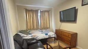 Une pièce de taille modeste comprenant un lit, un canapé en cuir noir, une commode en bois et un téléviseur fixé au mur.