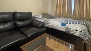 Une pièce comprenant un canapé en cuir noir, une table basse avec un plateau en verre, un lit recouvert d’une couvre-lit à motifs et une fenêtre avec des rideaux.