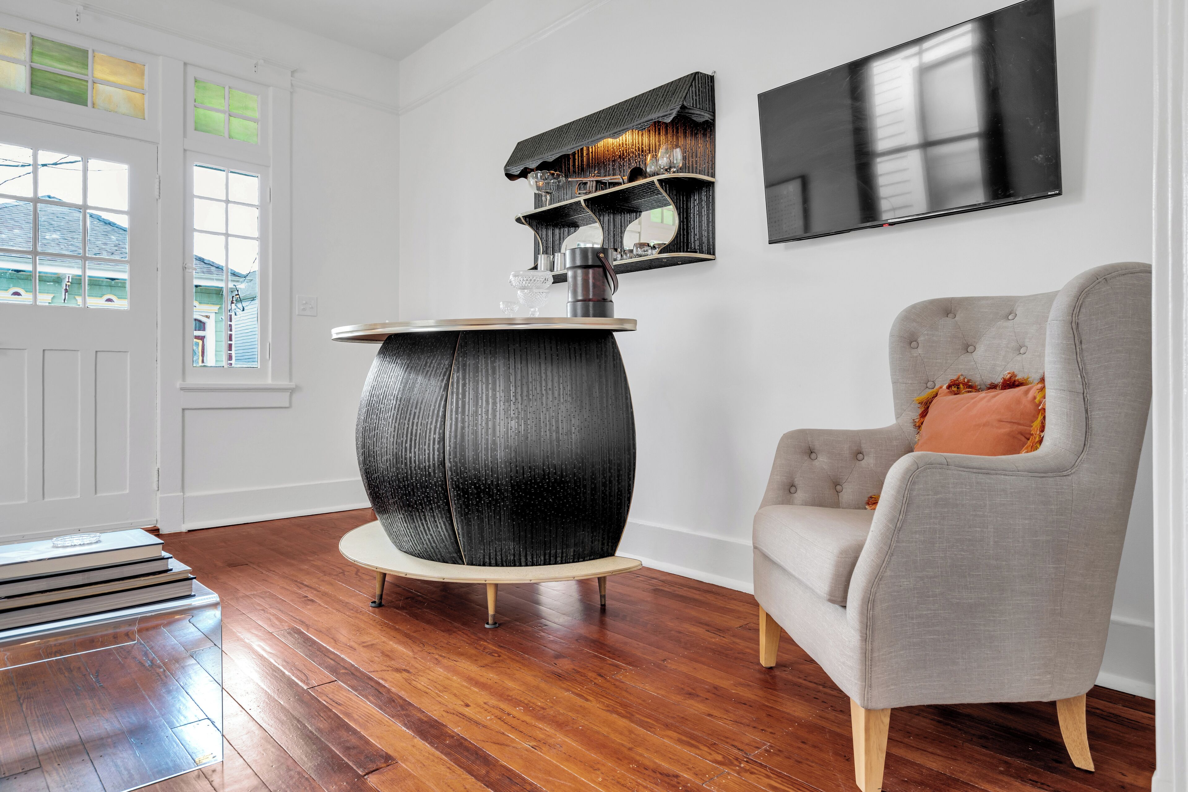 Une pièce avec un plancher en bois, une table ronde noire avec un plateau blanc, un fauteuil gris avec un coussin, une étagère murale ornée d’objets décoratifs et un téléviseur à écran plat.