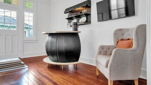 Una stanza con pavimento in legno, un tavolo rotondo nero con piano bianco, una poltrona grigia con cuscino, una mensola a parete con oggetti decorativi e un televisore a schermo piatto.