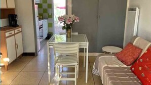 Een kleine kamer met een kitchenette, een eettafel met stoelen, een bank en een bijzettafeltje met een bloevas.