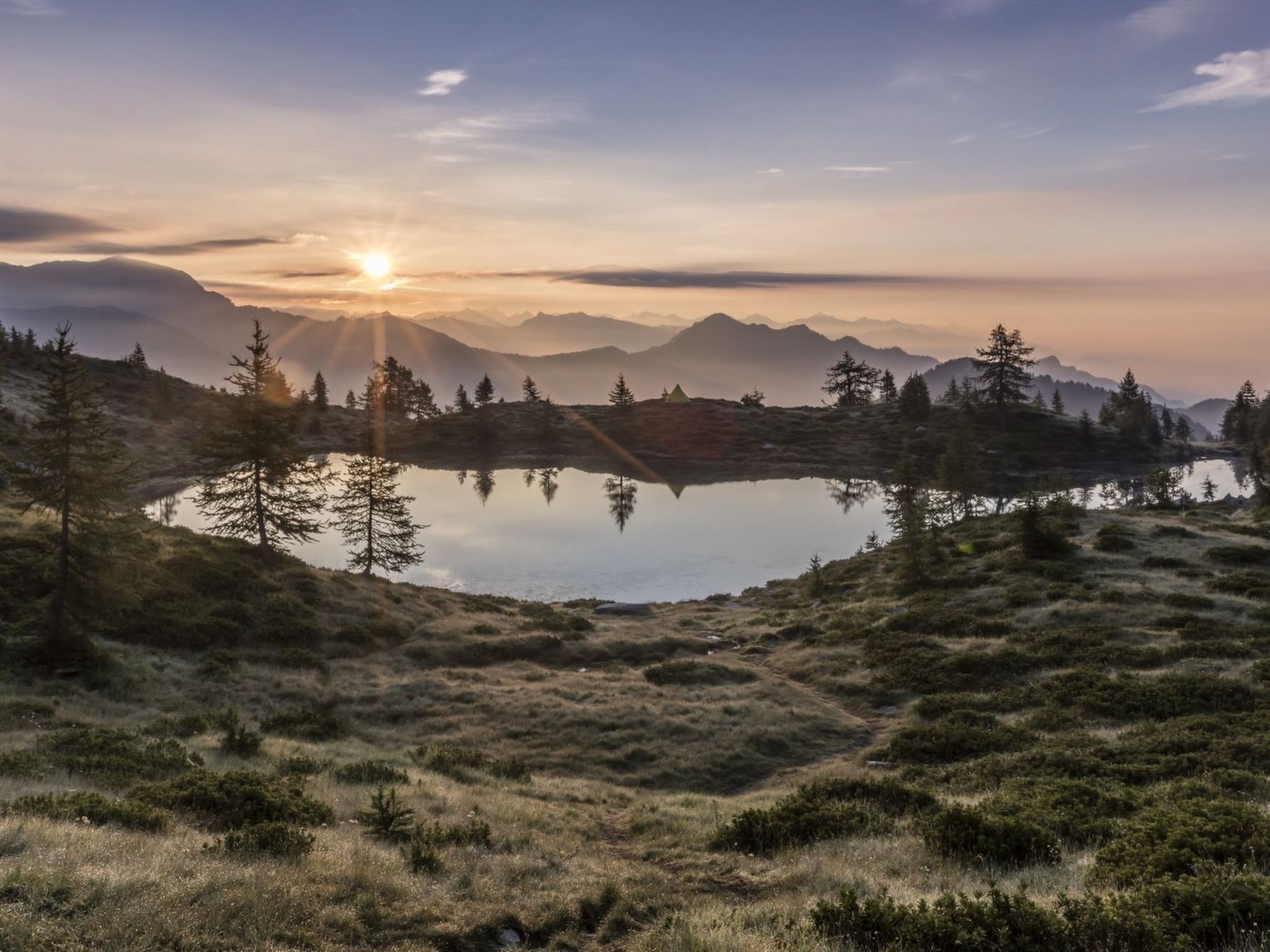 A mountainous landscape with a lake reflecting the sun, surrounded by trees and a clear sky.