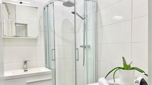 Un baño moderno con cabina de ducha de vidrio, tocador blanco con cajones, espejo y una pequeña planta en maceta.