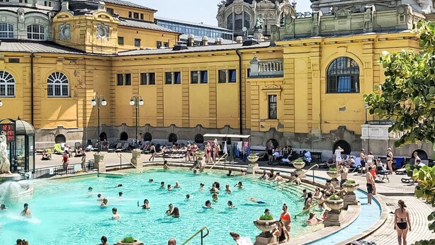 En allmän badplats med folk som simmar och solar, omgiven av historiska, gulmålade byggnader med kupolformade tak.