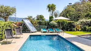 Una piscina rectangular con agua azul cristalina, rodeada por una solana empavedada. Hay sillones de descanso y una sombrilla, y la piscina está enmarcada por follaje y colinas.