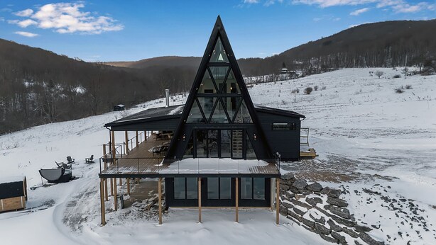Een modern, driehoekig gebouw met een glazen gevel, houten terras en een buiten zitgedeelte, gelegen in een besneeuwd landschap met heuvels op de achtergrond.