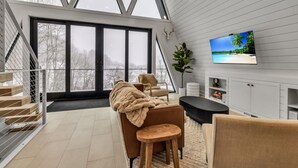 Een moderne woonkamer met een grote televisie, een open haard en zicht op een besneeerd landschap door grote ramen.