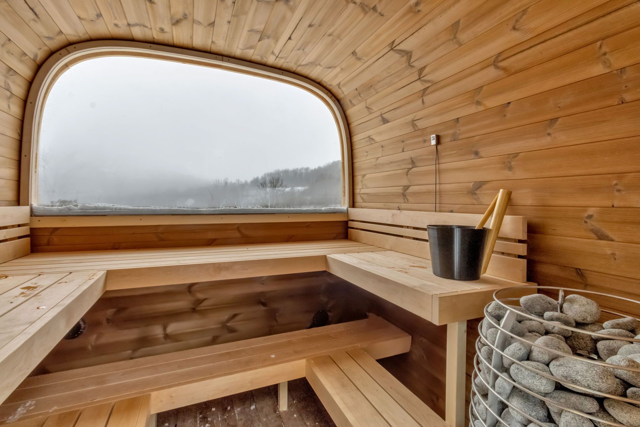 A wooden sauna with a bench, a window, a bucket, and a basket of stones.