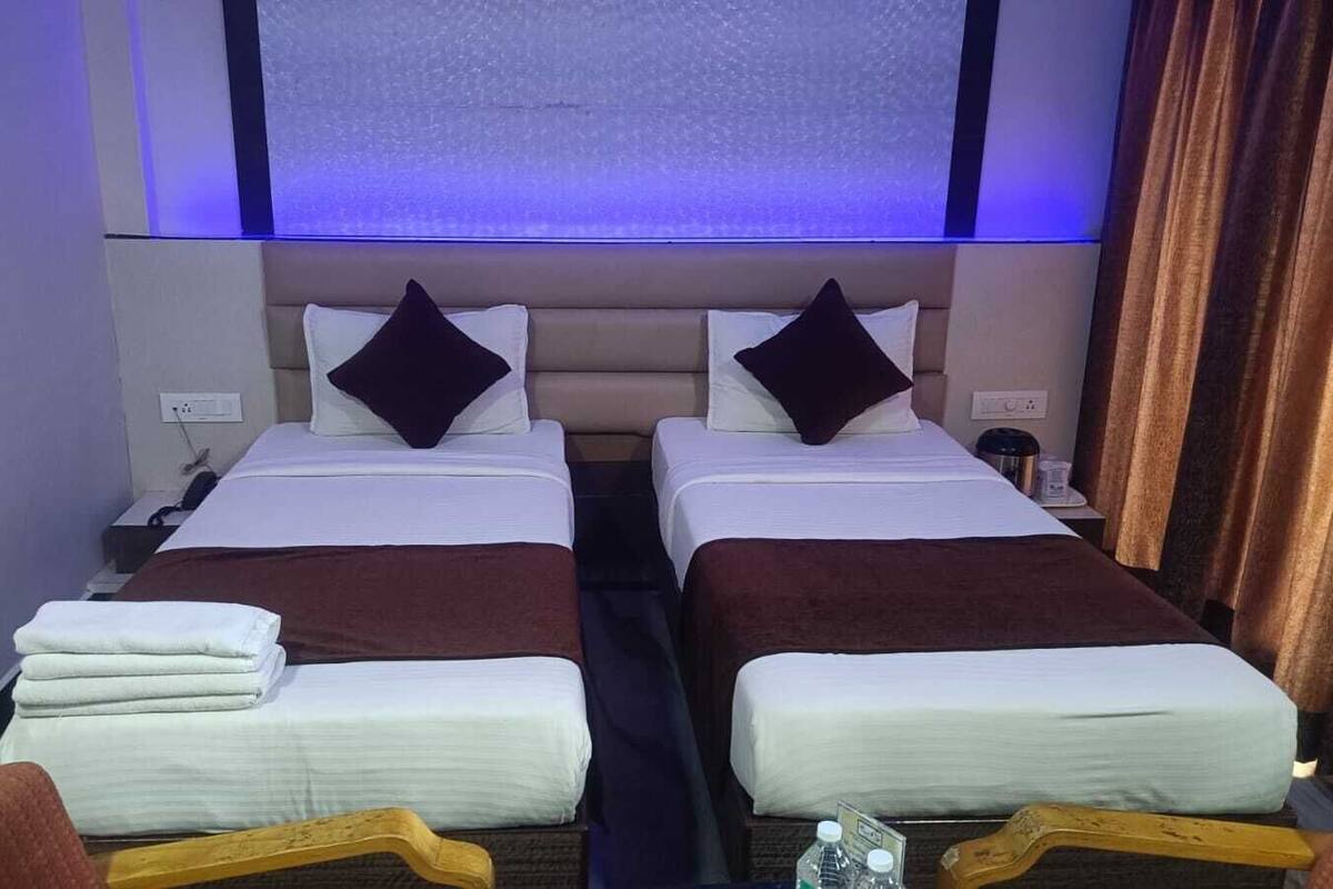 Habitación de hotel con dos camas, cada una con una almohada y una manta. Hay una mesita de noche con una lámpara y un pequeño escritorio con una botella y una caja de pañuelos.