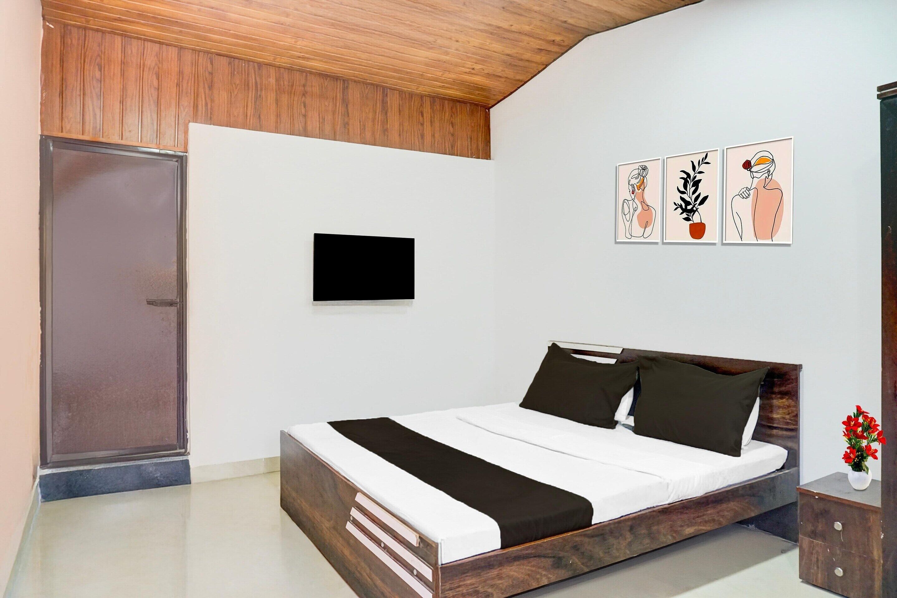 A bedroom with a wooden headboard, a flat-screen TV mounted on the wall, a bed with white and black bedding, and three framed artworks depicting a woman, a plant, and a man.