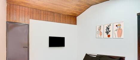 A bedroom with a wooden headboard, a flat-screen TV mounted on the wall, a bed with white and black bedding, and three framed artworks depicting a woman, a plant, and a man.