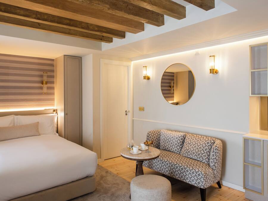 A hotel room with a bed, a round table with a cup and saucer, a patterned sofa, and a mirror on the wall.