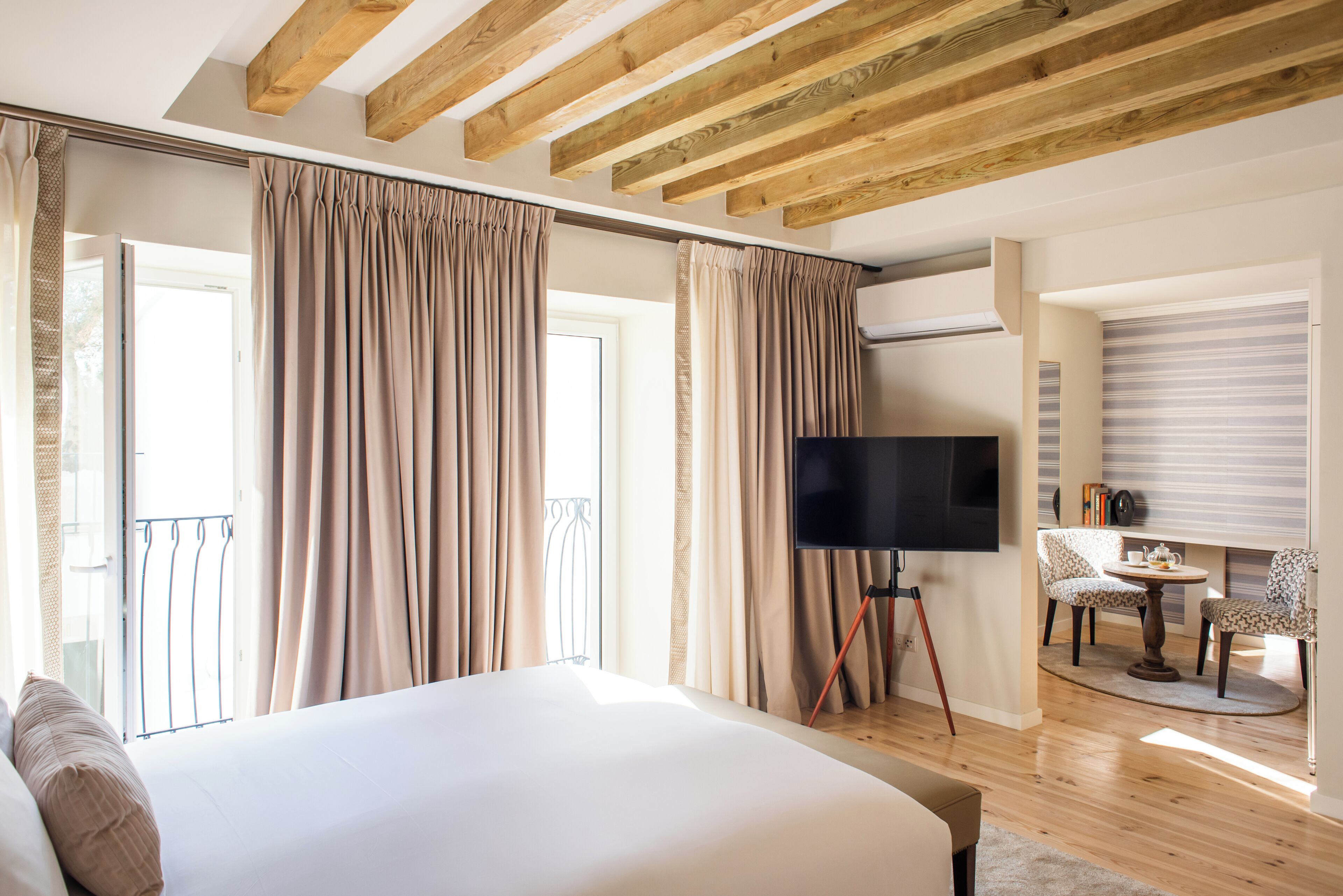 A bedroom with a large bed, a TV on a stand, a balcony with curtains, and a dining area with chairs.