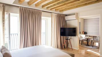 A bedroom with a large bed, a TV on a stand, a balcony with curtains, and a dining area with chairs.