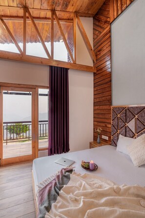 A bedroom with a wooden ceiling, a bed with a laptop, a vase with flowers, and a view of a balcony with a railing.