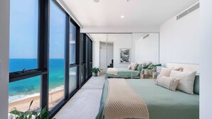 A modern bedroom with a large window overlooking a beach, a bed with a green bedspread, and a bedside table with a plant. - Elite Escape - Absolute Beachfront Luxury (Newcastle)