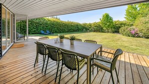Una terrazza esterna con tavolo e sedie in legno, una porta vetrata che dà su un giardino e vista su un prato ben curato e alberi.