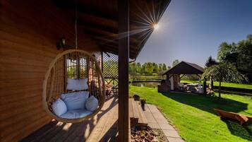 A wooden deck with a hanging chair, a covered seating area, and a view of a pond and garden.