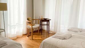 A hotel room with a wooden floor, a bed, a chair, a small table with a bowl and a glass, and sheer curtains.