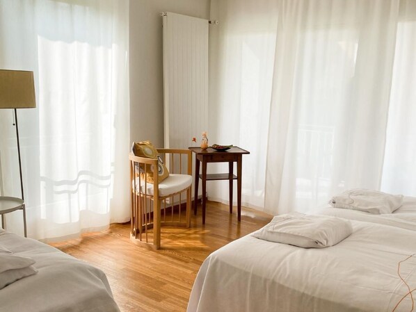 Quarto de hotel com piso de madeira, uma cama, uma cadeira, uma mesinha com uma tigela e um copo, e cortinas leves.