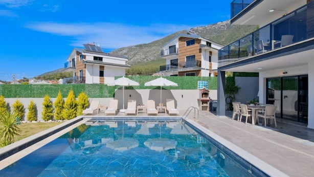 A modern pool area with lounge chairs and umbrellas, surrounded by contemporary buildings and a hillside backdrop.