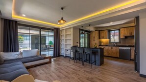 Ein modernes Wohnzimmer mit einer Couch und einem Essbereich mit Barhocker. Es gibt eine Küche mit Holzschränken und einem Fenster.