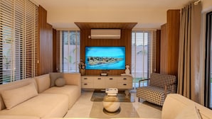 A modern living room with a large flat-screen TV, beige sofas, a patterned armchair, and wooden paneling.
