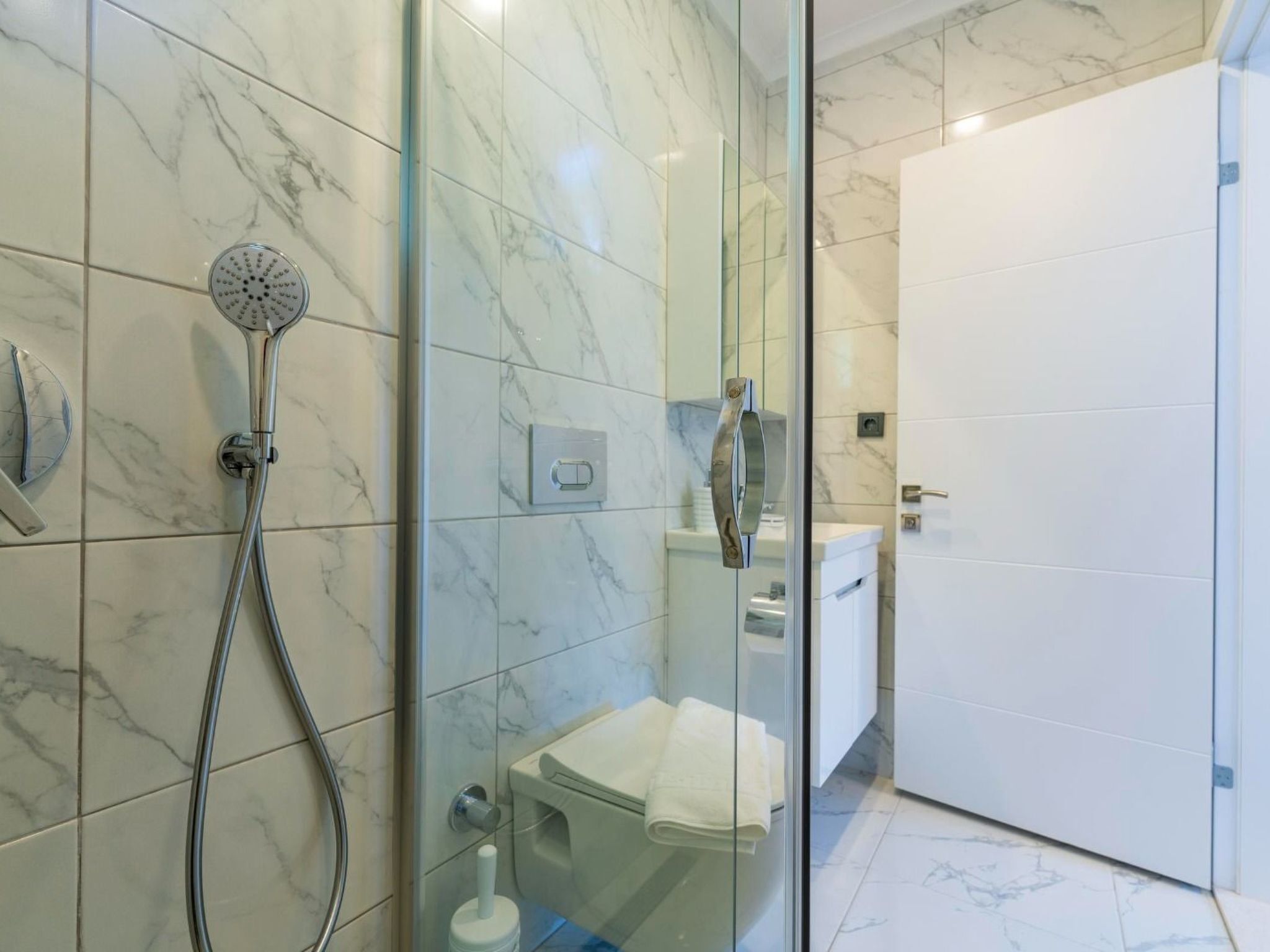 A modern bathroom with a glass shower enclosure, a handheld showerhead, and a toilet visible through the glass.