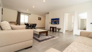 A modern living room with a sofa set, a coffee table, a dining table with chairs, and a flat-screen TV mounted on the wall.