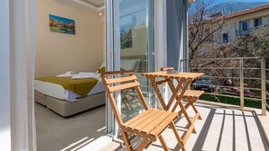 A balcony with a bed and wooden chairs, a painting on the wall, and a view of a building and trees.