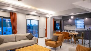 Un salón moderno con sofá, sillón y mesa de centro. Hay una zona de comedor con mesa y sillas. La habitación tiene ventanas grandes y vistas al exterior.