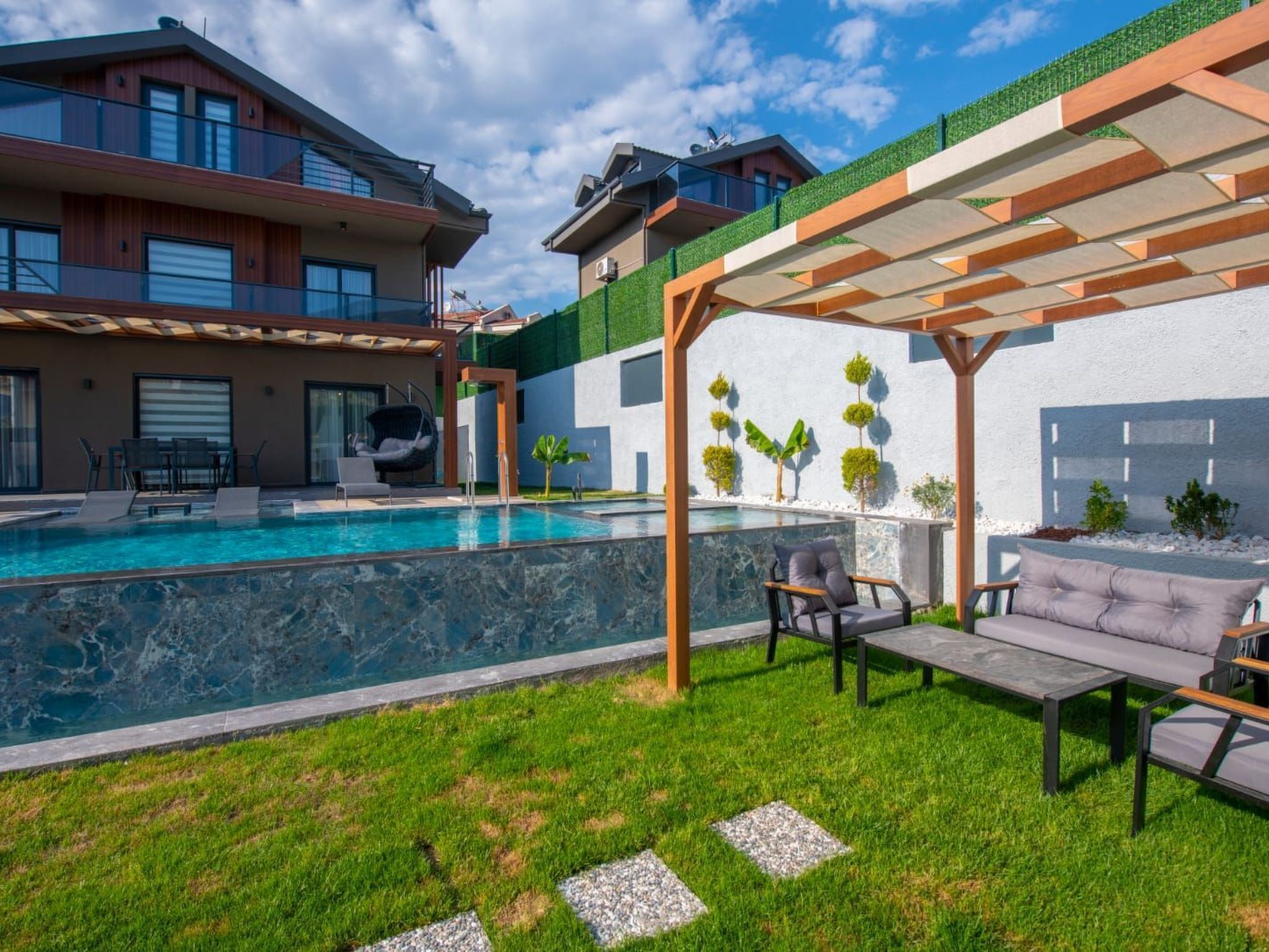 A modern outdoor pool area with a pergola, seating, and a view of a multi-story building.