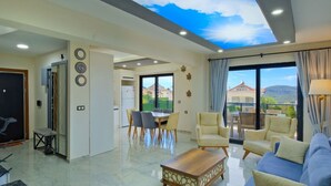 A spacious living room with a high ceiling, a dining area, and a view of the surrounding landscape.