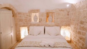 Un dormitorio con pared de piedra, una cama con sábanas blancas y dos lámparas fijadas a la pared.