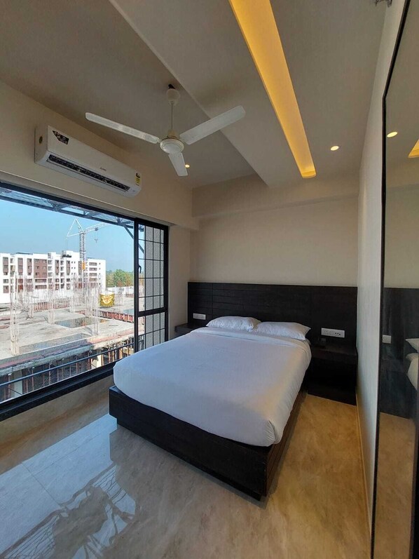 A modern bedroom with a large bed, a ceiling fan, and a view of a construction site through a large window. - The Forresta (Jalpaiguri)
